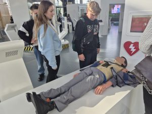 Uczniowie oglądają stanowisko edukacyjne poświęcone pierwszej pomocy, przy którym znajduje się fantom człowieka oraz ekran z symbolem defibrylatora AED.