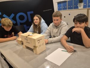 Czworo uczniów siedzi przy stole z drewnianymi skrzynkami zabezpieczonymi kłódkami podczas warsztatów z łamania szyfrów w Centrum Nauki Experyment.