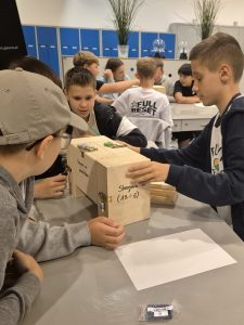 Uczniowie pracują w grupach nad otwieraniem drewnianych skrzynek z zamkami szyfrowymi, rozwiązując zagadki podczas warsztatów „Złam Enigmę!”.