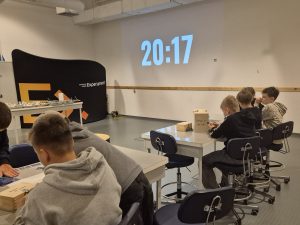 Uczniowie pracują w zespołach nad zagadkami matematycznymi w sali warsztatowej Centrum Nauki Experyment. Na ścianie wyświetlany jest zegar odliczający czas.