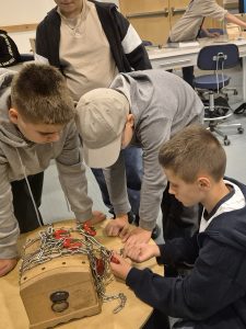 Uczniowie wspólnie próbują otworzyć drewnianą skrzynię zabezpieczoną łańcuchami i kłódkami podczas warsztatów „Złam Enigmę!” w Centrum Nauki Experyment w Gdyni.