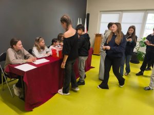 Uczniowie oddają głosy w wyborach do Samorządu Uczniowskiego. Przy stole przykrytym bordowym obrusem siedzą członkinie komisji wyborczej, a uczniowie podchodzą do czerwonej urny i pobierają karty do głosowania.