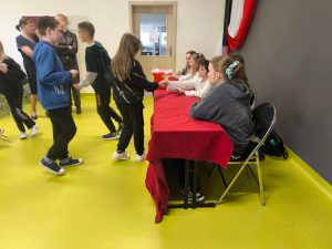Uczniowie uczestniczą w szkolnych wyborach, podchodząc kolejno do komisji wyborczej. Przy stole z czerwoną urną siedzą uczennice odpowiedzialne za przeprowadzenie głosowania. W tle widoczni są pozostali uczniowie oraz nauczyciele.
