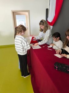 Uczennica z komisji wyborczej sprawdza dokumenty i przekazuje kartę do głosowania młodszemu uczniowi. Przy stole przykrytym bordowym obrusem pozostałe członkinie komisji przygotowują karty i dokumentację wyborczą.
