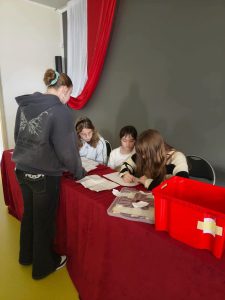 Uczennice komisji wyborczej przeglądają karty do głosowania i zapisują wyniki na przygotowanych arkuszach. Na stole przykrytym bordowym obrusem znajdują się dokumenty wyborcze, przezroczysty pojemnik z kartami oraz czerwona urna.