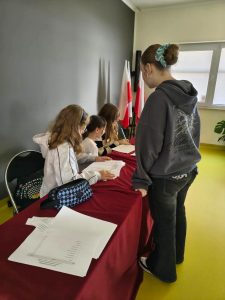 Członkinie komisji wyborczej sprawdzają listy i dokumentację po zakończonym głosowaniu. Przy stole stoją i siedzą uczennice analizujące wyniki wyborów. W tle widoczne są biało-czerwone flagi.