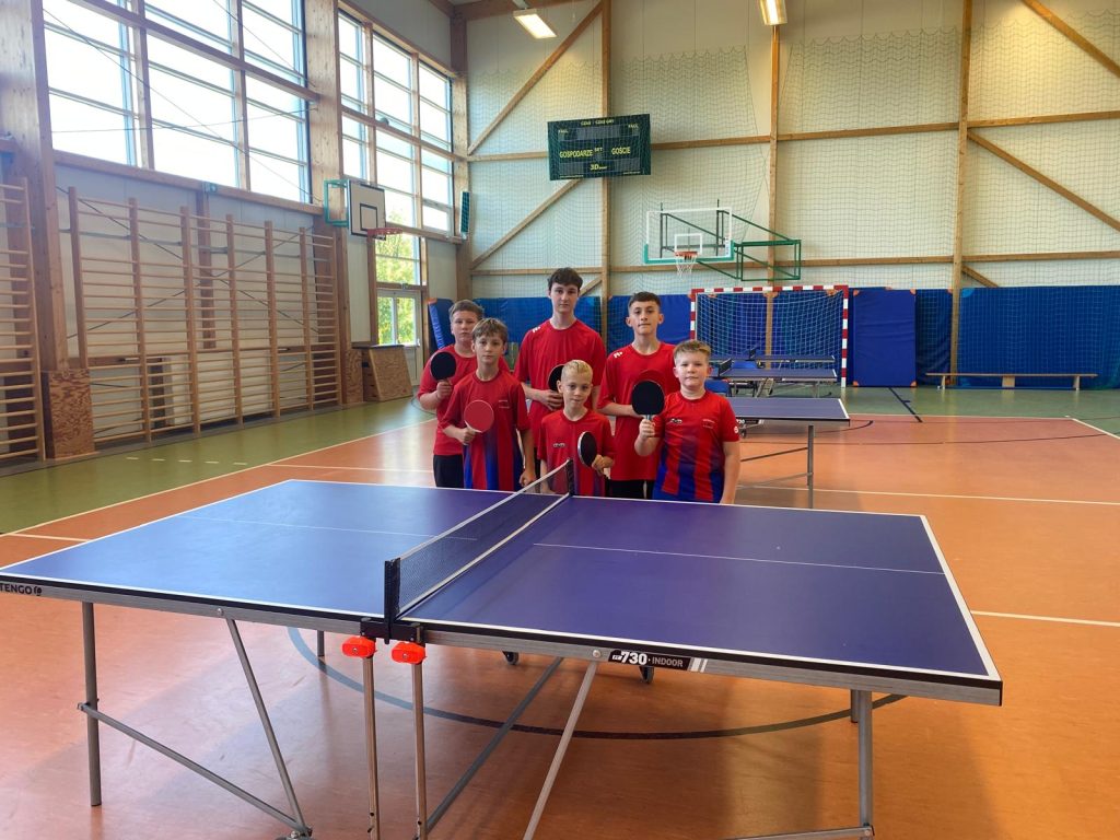 Sześciu uczniów Szkoły Podstawowej w Ostaszewie w czerwonych koszulkach sportowych pozuje z rakietkami do tenisa stołowego za stołem pingpongowym w hali sportowej podczas Drużynowych Mistrzostw Gminy Łysomice.