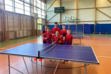Sześciu uczniów Szkoły Podstawowej w Ostaszewie w czerwonych koszulkach sportowych pozuje z rakietkami do tenisa stołowego za stołem pingpongowym w hali sportowej podczas Drużynowych Mistrzostw Gminy Łysomice.