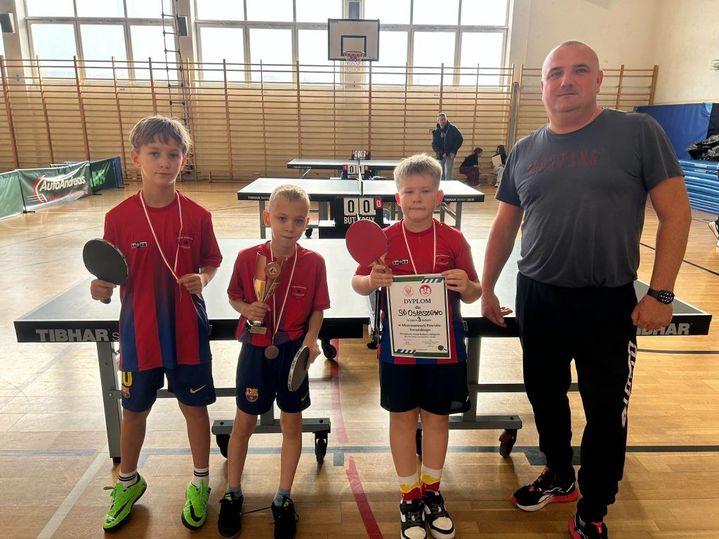 Trzej uczniowie SP Ostaszewo w czerwono-granatowych strojach sportowych, z medalami na szyjach, pozują do zdjęcia wraz z opiekunem na sali gimnastycznej. Uczniowie trzymają rakietki do tenisa stołowego, puchar oraz dyplom za zajęcie III miejsca w Drużynowych Mistrzostwach Powiatu Toruńskiego w Tenisie Stołowym w kategorii Igrzyska Dzieci. W tle widoczne są stoły do tenisa stołowego.